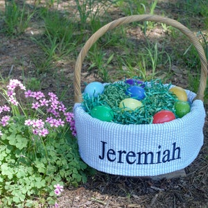 May include: An Easter basket with a woven handle, filled with green paper grass and colorful eggs. The basket's blue and white striped fabric is embroidered with the name "Jeremiah". The basket is set outdoors near pink flowers.