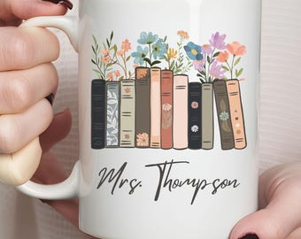 May include: White ceramic mug featuring a floral and book design. The design includes a row of books with colorful flowers on top. The name "Mrs. Thompson" is written in script below the books.