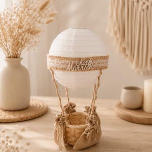 May include: A decorative hot air balloon lamp featuring a white paper lantern, a woven basket, and a burlap basket. The lamp has a wooden band with the words "Just Married". Ideal for wedding or home decor.