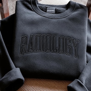 May include: A black crewneck sweatshirt featuring the word RADIOLOGY embroidered in a matching black thread across the chest in a collegiate-style font. The garment has long sleeves with cuffed ends and a small interior neck label.