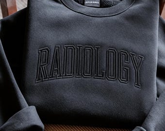 May include: A black crewneck sweatshirt featuring the word RADIOLOGY embroidered in a matching black thread across the chest in a collegiate-style font. The garment has long sleeves with cuffed ends and a small interior neck label.