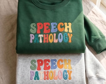 Può includere: Una felpa girocollo verde scuro e una felpa girocollo grigio chiaro, entrambe con la scritta SPEECH PATHOLOGY ricamata in un carattere ondulato multicolore con lettere nei toni del verde, rosso, blu, arancione, giallo e rosa.