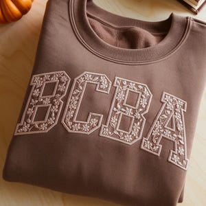May include: A brown crewneck sweatshirt with the embroidered letters "BCBA" in a floral design. The sweatshirt is folded on a light wood surface, with a small pumpkin and a book in the background.