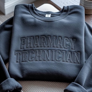 May include: A dark charcoal gray crewneck sweatshirt featuring the words PHARMACY TECHNICIAN embroidered in a matching dark thread across the chest in a bold, collegiate-style block font. The garment has long sleeves and a ribbed neckline.