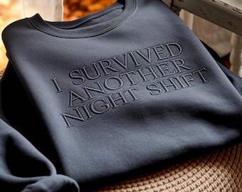 Op de afbeelding: Een donker antracietgrijze sweater met ronde hals en de geborduurde tekst I SURVIVED ANOTHER NIGHT SHIFT in een bijpassende kleur garen. Het kledingstuk heeft lange mouwen, een geribbelde halslijn en een klassiek pullover-ontwerp.