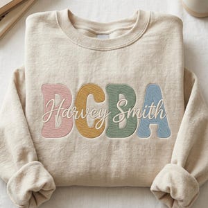 May include: A beige crewneck sweatshirt featuring large, embroidered block letters in muted pink, mustard yellow, sage green, and dusty blue. Overlaid across the centre is the name Harvey Smith in white cursive script embroidery.