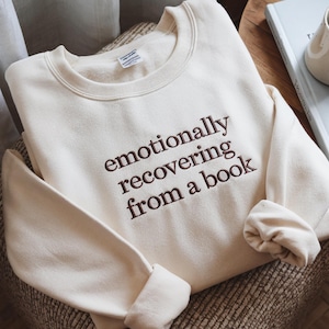 May include: A cream-colored crewneck sweatshirt featuring dark brown embroidered text on the chest that reads, "emotionally recovering from a book." The garment has long sleeves with ribbed cuffs and a ribbed neckline, showcasing a soft fabric texture.