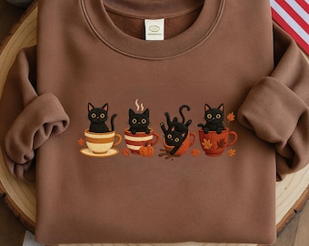 May include: A brown sweatshirt with an embroidered design of four black cats in teacups. The cats are in different poses, with autumn leaves and pumpkins. The sweatshirt has a crew neck and long sleeves.