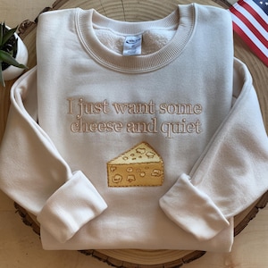 Puede incluir: Una sudadera de cuello redondo color crema con el texto bordado "I just want some cheese and quiet" en hilo marrón, situado sobre un trozo de queso suizo amarillo bordado con agujeros visibles y contornos de color marrón.