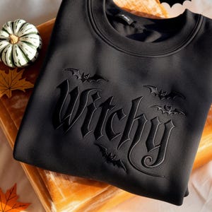 May include: A black long-sleeve sweatshirt featuring the word Witchy embossed in a gothic font across the chest. The design includes three small bat silhouettes integrated around the lettering, with a crew neckline and a solid black fabric finish.