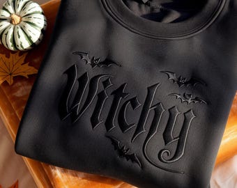 May include: A black long-sleeve sweatshirt featuring the word Witchy embossed in a gothic font across the chest. The design includes three small bat silhouettes integrated around the lettering, with a crew neckline and a solid black fabric finish.