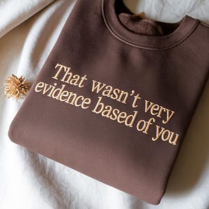 May include: A brown sweatshirt with the embroidered text "That wasn't very evidence based of you" in a light tan color. The sweatshirt is folded and resting on a white surface.