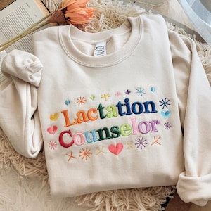May include: A cream-colored crewneck sweatshirt featuring colorful embroidered text that reads Lactation Counselor. The design includes various small embroidered starbursts and heart shapes in shades of pink, blue, green, orange, and purple.