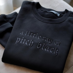 May include: A black sweatshirt with the embroidered phrase "a little ray of pitch black" in a gothic font. The crew neck sweatshirt is folded neatly on a light-colored surface.