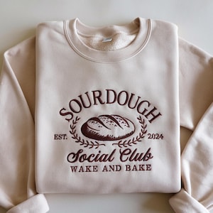 May include: A beige sweatshirt with the embroidered text "SOURDOUGH SOCIAL CLUB WAKE AND BAKE" in brown. The design includes a bread loaf illustration and the year "2024". The sweatshirt has a crew neck and long sleeves.