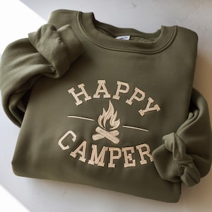 May include: An olive green sweatshirt with the embroidered text "HAPPY CAMPER" above a campfire graphic. The sweatshirt has a crew neck and long sleeves. The design is in a cream color, creating a contrast against the green fabric.