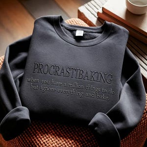 May include: A dark charcoal gray crewneck sweatshirt featuring the embroidered text "PROCRASTIBAKING" in a bold font, followed by the smaller embroidered text "when you have a million things to do but ignore everything and bake,".