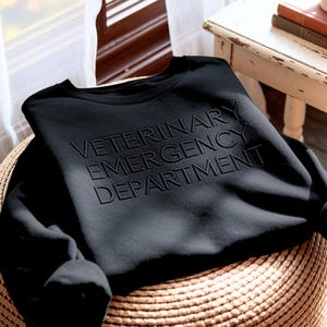 May include: A black crewneck sweatshirt featuring the embossed text VETERINARY EMERGENCY DEPARTMENT centered on the chest. The garment has long sleeves and a ribbed neckline, showcasing a monochromatic design with raised lettering in the same color.