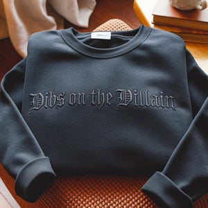 May include: A dark gray sweatshirt with the phrase "Dibs on the Villain" embroidered in a gothic font across the chest. The sweatshirt is folded neatly on a woven surface, with the sleeves cuffed.
