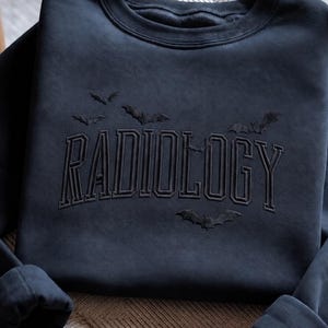May include: A black crewneck sweatshirt featuring the word RADIOLOGY embroidered in a matching black thread across the chest. The design includes several small, embroidered bat silhouettes flying above and below the text in black thread.