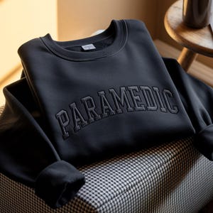 May include: A black crewneck sweatshirt featuring the word PARAMEDIC embroidered in a matching black, arched, block-style font across the chest. The garment has long sleeves with ribbed cuffs and a ribbed neckline, showcasing a clean, solid design.