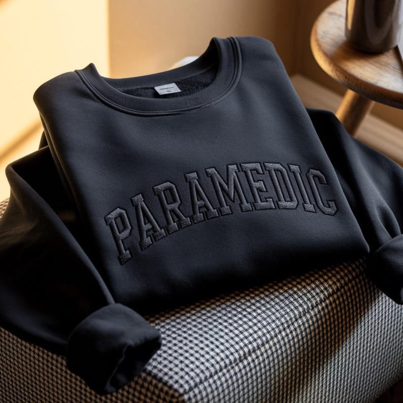 May include: A black crewneck sweatshirt featuring the word PARAMEDIC embroidered in a matching black, arched, block-style font across the chest. The garment has long sleeves with ribbed cuffs and a ribbed neckline, showcasing a clean, solid design.
