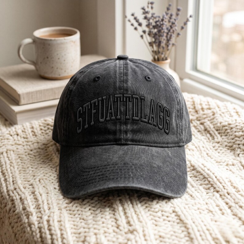 K&ouml;nnte beinhalten: Eine dunkelgraue Baseballkappe mit dem gestickten Text "STFUATTDLAGG" auf der Vorderseite. Die Kappe liegt auf einer cremefarbenen, strukturierten Oberfl&auml;che. Eine Keramiktasse und B&uuml;cher sind im Hintergrund zu sehen.