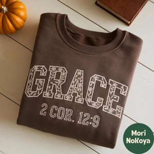 May include: A dark brown crewneck sweatshirt featuring the word GRACE embroidered in large white block letters filled with a delicate floral vine pattern. Below the word, the text 2 COR. 12:9 is embroidered in a simple white font.