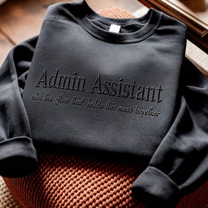 May include: Dark gray sweatshirt with the words "Admin Assistant" embossed on the front, along with the phrase "aka the glue that holds this mess together." The sweatshirt is folded on a patterned surface.