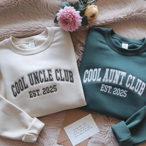 May include: Two crewneck sweatshirts, one cream and one green, with the text "COOL UNCLE CLUB EST. 2025" and "COOL AUNT CLUB EST. 2025" in a collegiate font. A pink and yellow flower arrangement and a card are in the background.