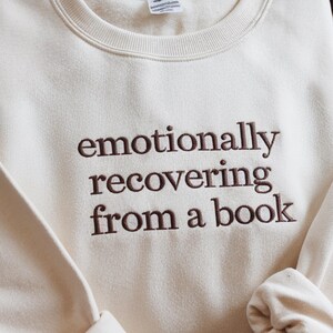 May include: A cream-coloured crewneck sweatshirt featuring dark brown embroidered text on the chest that reads, "emotionally recovering from a book." The garment has a ribbed neckline and long sleeves, showcasing a clean and simple design.