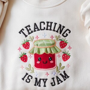May include: Cream-colored sweatshirt with the embroidered phrase "TEACHING IS MY JAM." The design features a smiling red jam jar with a green lid, surrounded by strawberries, leaves, and stars. A cute and playful design.