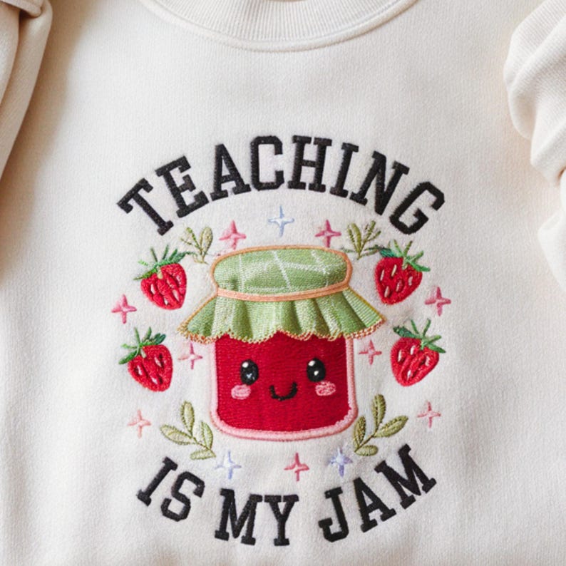 May include: Cream-colored sweatshirt with the embroidered phrase "TEACHING IS MY JAM." The design features a smiling red jam jar with a green lid, surrounded by strawberries, leaves, and stars. A cute and playful design.