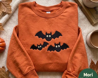 May include: A burnt orange long-sleeve sweatshirt featuring an embroidered design of three black bats with large white eyes. The garment has a crew neckline, ribbed cuffs, and a ribbed hem, showcasing a clean, solid-colored fabric construction.