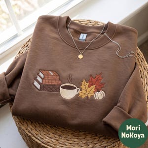Könnte beinhalten: Ein braunes Sweatshirt mit Rundhalsausschnitt und einer Stickerei, die einen Stapel aus drei Büchern, eine dampfende Kaffeetasse, zwei herbstliche Blätter in Gelb und Rostrot sowie einen kleinen weißen Kürbis zeigt. Eine goldfarbene Kette liegt am Ausschnitt.