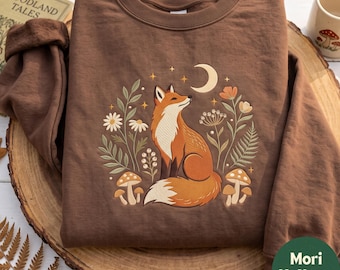 May include: A brown crewneck sweatshirt featuring a central graphic of a fox sitting among flowers, mushrooms, and a crescent moon. The design includes white daisies, green foliage, and orange blossoms, with small yellow stars scattered around the fox.