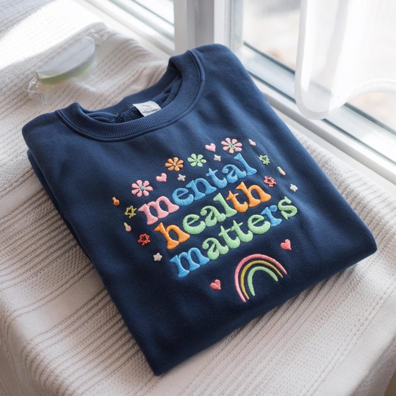 May include: A navy blue sweatshirt with the embroidered phrase "mental health matters" in colorful letters. The design includes flowers, hearts, stars, and a rainbow. The sweatshirt is folded on a white surface near a window.