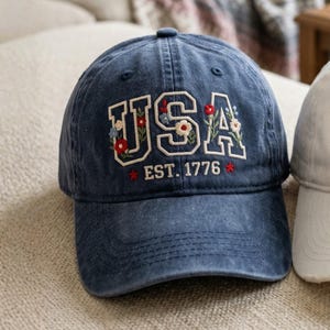 May include: A navy blue baseball cap with the word "USA" embroidered in white, decorated with floral accents. The text "EST. 1776" is below the word "USA". The cap is sitting on a beige surface.
