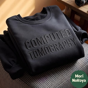 May include: A black crewneck sweatshirt featuring the words COMPUTED TOMOGRAPHY embossed in a matching black, raised font across the chest. The garment has long sleeves with ribbed cuffs and a ribbed neckline, presented in a clean, folded style.
