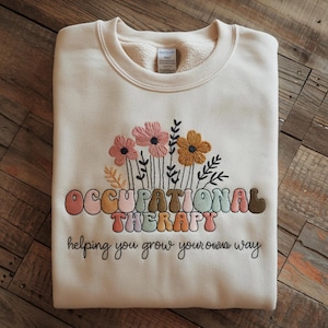 May include: Cream-colored sweatshirt with embroidered flowers and the words "OCCUPATIONAL THERAPY" in a retro font. Below the text is the phrase "helping you grow your own way" in a script font. The sweatshirt is folded on a wooden surface.