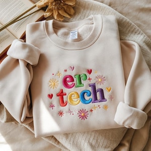 May include: Cream-colored sweatshirt with the embroidered text "er tech" in rainbow colors, surrounded by floral and heart designs. The long sleeves are cuffed. The sweatshirt is folded on a neutral-toned surface.