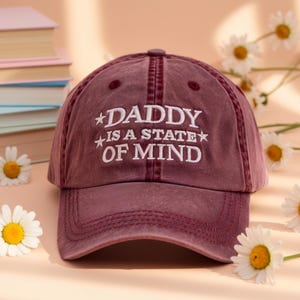 May include: A maroon baseball cap with the text "DADDY IS A STATE OF MIND" embroidered in white. The cap has a curved brim and a classic six-panel design. The hat is surrounded by daisies.