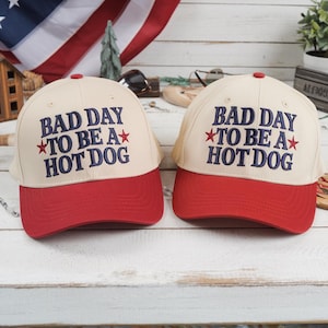 Op de afbeelding: Twee baseballpetten met een cr&egrave;mekleurige kroon en een rode klep. De petten hebben de geborduurde tekst "BAD DAY TO BE A HOT DOG" in marineblauw met rode steraccenten. Ze liggen op een wit houten oppervlak.