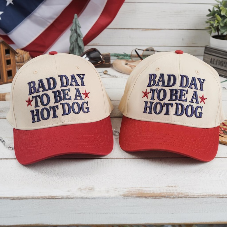 Op de afbeelding: Twee baseballpetten met een cr&egrave;mekleurige kroon en een rode klep. De petten hebben de geborduurde tekst "BAD DAY TO BE A HOT DOG" in marineblauw met rode steraccenten. Ze liggen op een wit houten oppervlak.