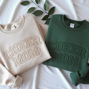 May include: Two crewneck sweatshirts, one in beige and one in dark green, each featuring the text CHEER MOM embroidered in a collegiate font across the chest. The sweatshirts have long sleeves, ribbed necklines, and a soft, fleece-lined interior.