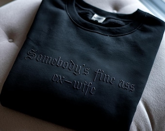 May include: Black crewneck sweatshirt with the phrase "Somebody's fine ass ex-wife" embroidered in a gothic font. The sweatshirt is folded neatly on a light-colored surface.