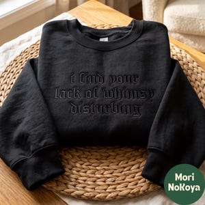 May include: A black crewneck sweatshirt featuring the embroidered text "i find your lack of whimsy disturbing" in a matching black thread. The garment has long sleeves with ribbed cuffs and a ribbed neckline, showcasing a monochromatic design.