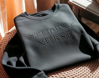 May include: A dark gray sweatshirt with the words "STUDENT NURSE" embroidered across the chest. The sweatshirt is folded on a woven, round, tan-colored cushion. The background includes a wooden side table and a beige curtain.