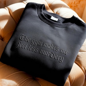 May include: Black crew neck sweatshirt with the phrase "The gym makes me feel less murdery" in an embossed gothic font. The sweatshirt is made of a soft material and is displayed on a beige upholstered surface.