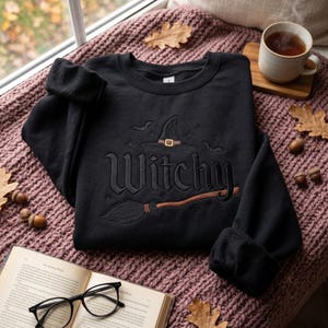 May include: A black long-sleeve sweatshirt featuring an embroidered design on the chest. The embroidery includes a witch hat, a broomstick, small bats, and the word "Witchy" in a stylized font. The sweatshirt has a crew neckline and ribbed cuffs.
